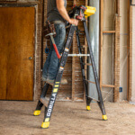 Worker on a black Gorilla ladder with yellow accents, inside a construction area.