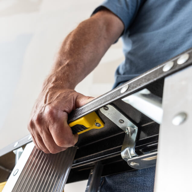 Hand gripping a silver ladder with a yellow lock mechanism; focus on ladder detail.