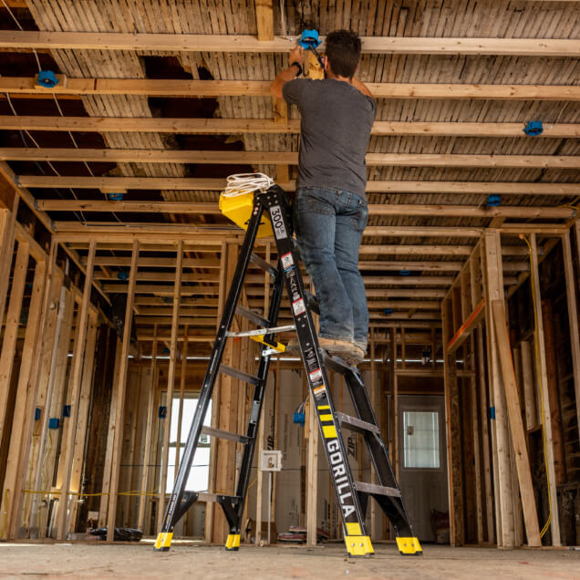Person standing on black Gorilla ladder adjusting ceiling wiring in unfinished room.
