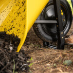 Yellow wheelbarrow with black wheel on dirt, loaded with dark mulch.