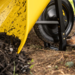 Yellow wheelbarrow with black wheel on dirt, loaded with dark mulch.