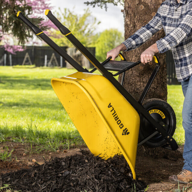 Yellow Gorilla wheelbarrow with flat-free tire, dumping mulch near a tree.
