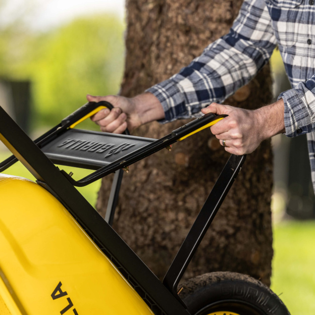 Person holding a yellow Gorilla wheelbarrow handle with "GORILLA" text, blurred tree background.