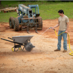 Person using Gorilla hose to spray a gray Gorilla wheelbarrow near machinery.